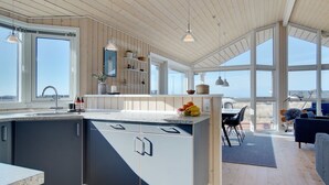 A modern kitchen with a large island, wooden ceiling, and large windows offering a view of the beach.
