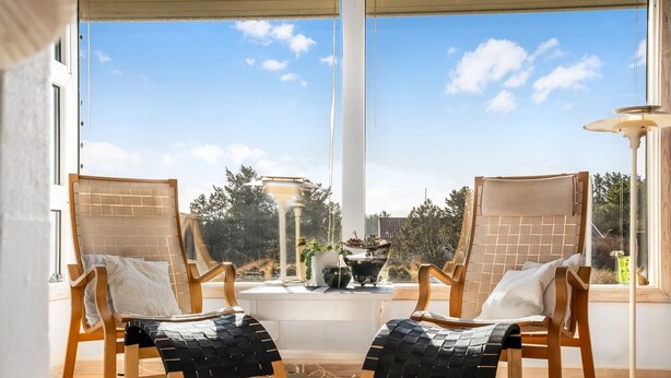 Quarto com duas cadeiras de madeira com almofadas pretas, uma mesa branca e vista para árvores e céu através de grandes janelas.
