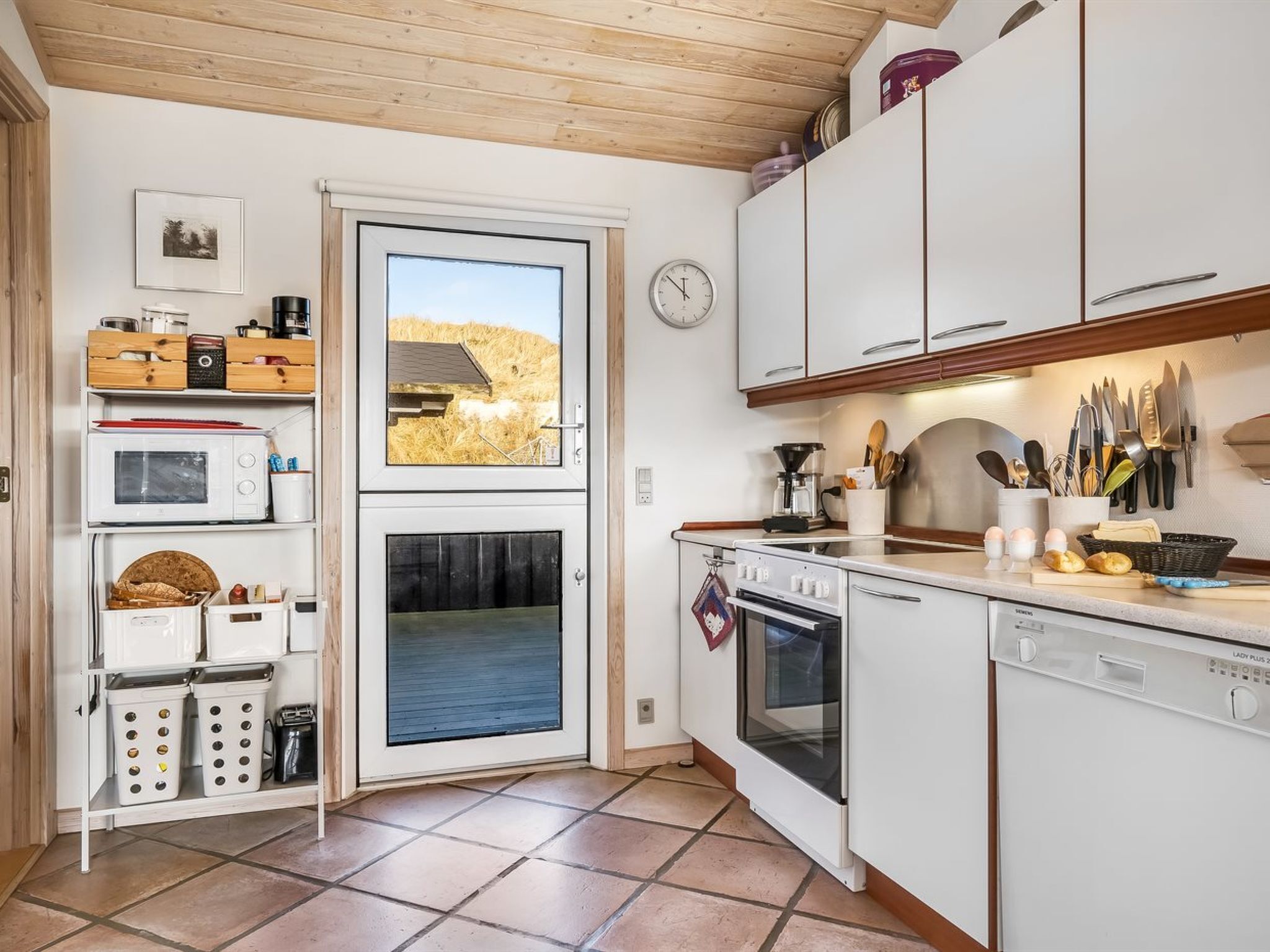 Cocina compacta con microondas, horno y lavavajillas. Hay una ventana con vistas a un edificio y un reloj en la pared.