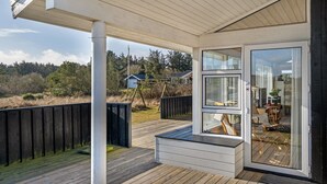 Área externa coberta com deck de madeira, porta de vidro que dá para um ambiente interno e vista para uma área residencial com árvores e uma casa ao fundo.