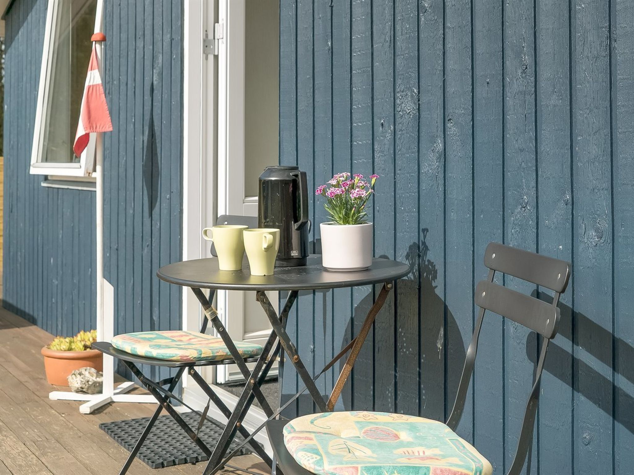 A small outdoor seating area with a table, two chairs, a coffee pot, and a potted plant.