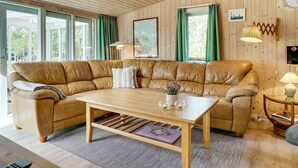 A cozy living room with a large leather sectional sofa, a wooden coffee table, and a view through large windows with green curtains.