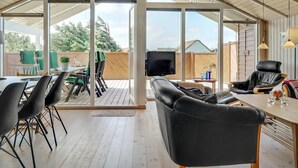 Um quarto espaçoso com uma grande porta de vidro deslizante que dá para um pátio externo. No interior, há uma área de estar confortável com um sofá e cadeiras, uma mesa de jantar e uma televisão de tela plana fixada na parede.