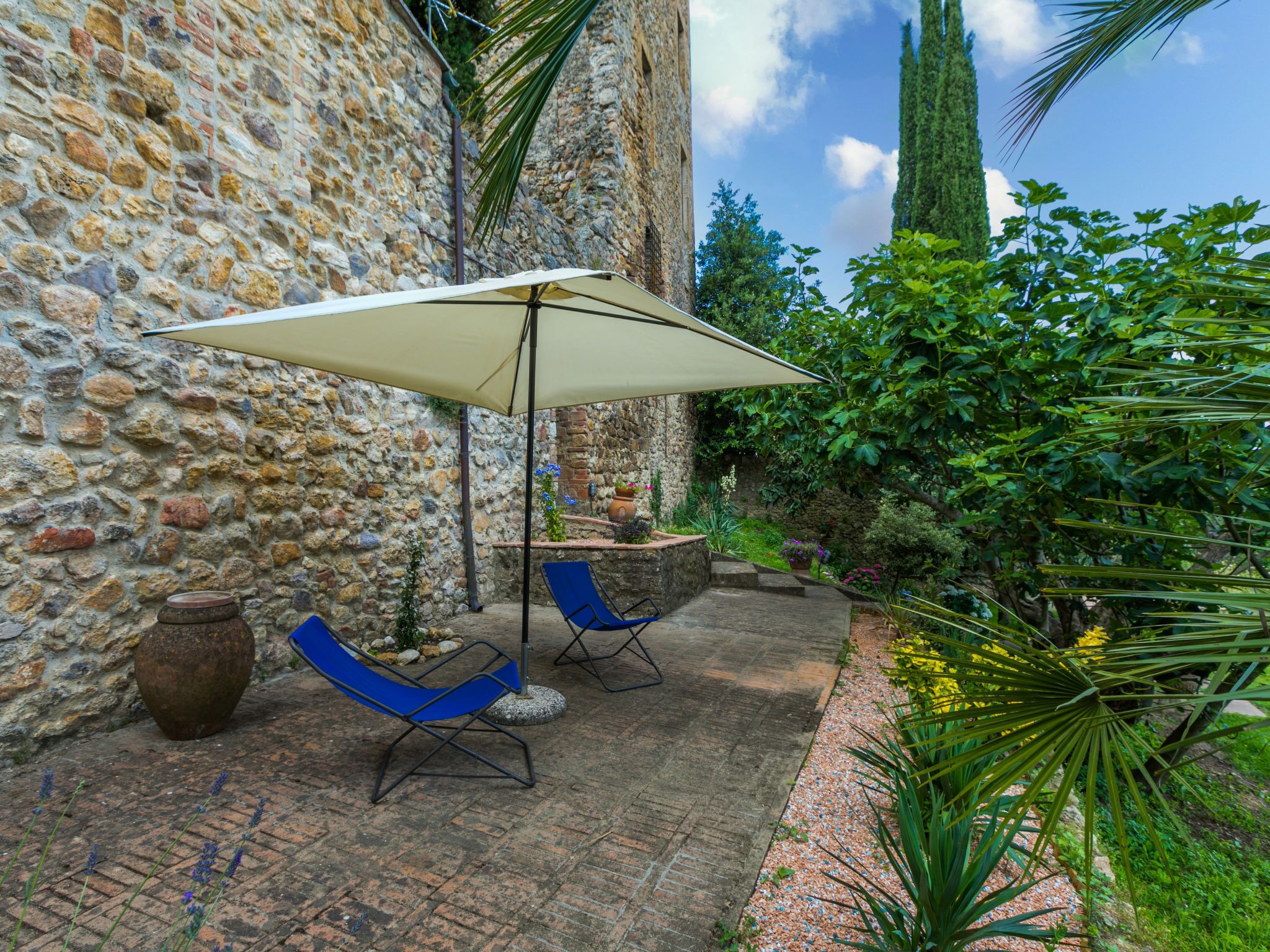 Eine Terrasse im Freien mit Steinmauern, zwei blauen Stühlen, einem großen Sonnenschirm und Kübelpflanzen.