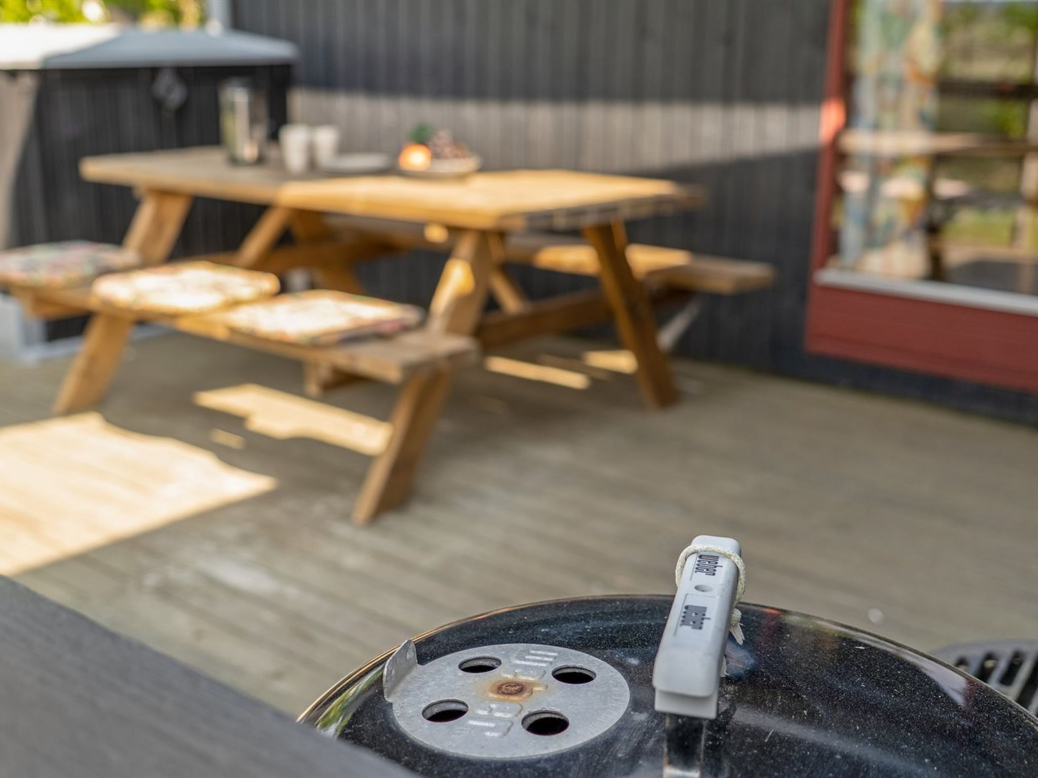 Una parrilla con termómetro sobre una terraza de madera, con mesa y sillas.