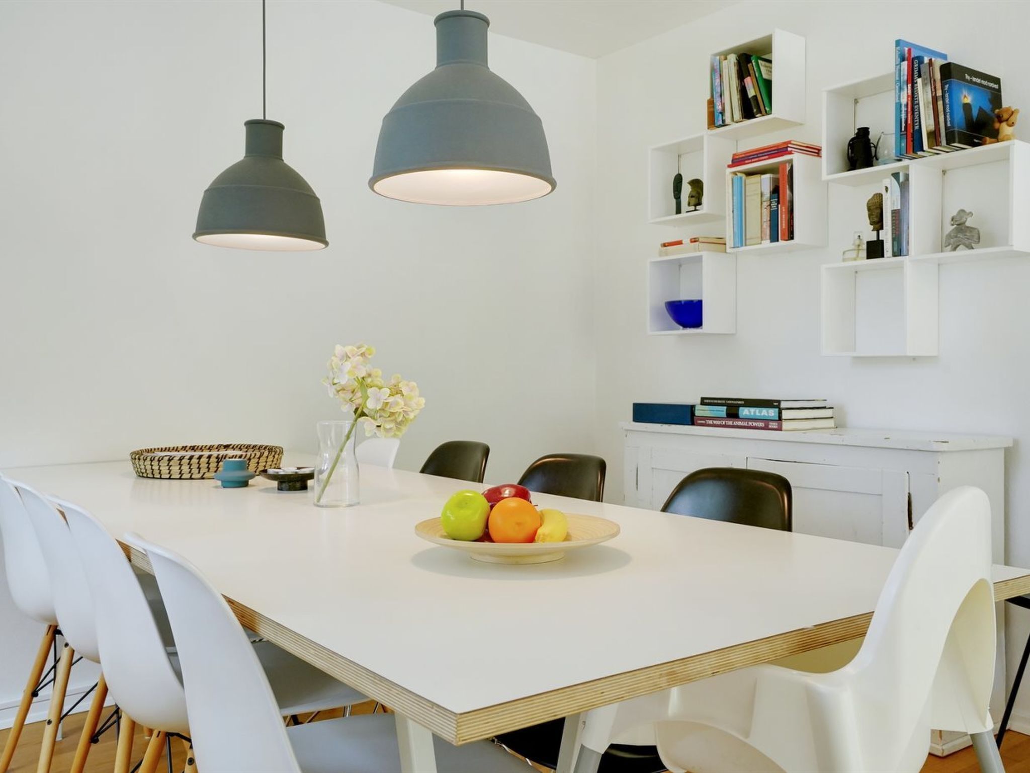 Een moderne eetruimte met een witte tafel, twee hanglampen, een fruitschaal en een boekenkast met boeken en decoratieve objecten.