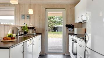 A modern kitchen with white cabinets, a stainless steel refrigerator, and a black countertop. There is a door leading to an outdoor area with a view of greenery.