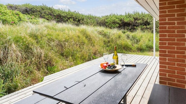 A wooden table with a bottle of wine, glasses, and a plate of fruit on an outdoor deck with a view of a grassy landscape. - "Annemia" - 200m from the sea by Interhome (Thisted)
