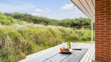 A wooden table with a bottle of wine, glasses, and a plate of fruit on an outdoor deck with a view of a grassy landscape.