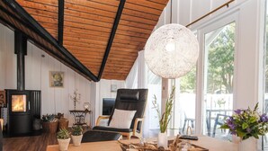 Un salon chaleureux avec une cheminée, un fauteuil confortable, une table en bois sur laquelle est posé un plateau, et une grande fenêtre donnant sur la forêt.