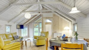 Ein geräumiger Raum mit hoher Decke, Holzbalken, einem gelben Sofa, einem gelben Sessel, einem Holzesstisch mit gemustertem Tischläufer und einem an der Wand montierten Fernseher.