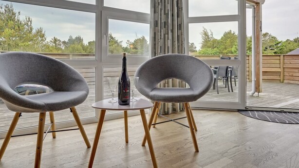 Een moderne buitenzitplaats met twee grijze stoelen en een bijzettafeltje, een fles wijn en uitzicht op het omliggende landschap door grote ramen.