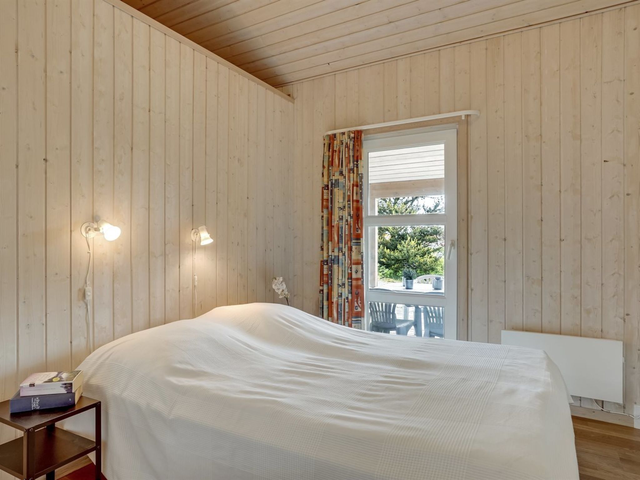Een slaapkamer met een bed, een nachtkastje, twee wandlampen, een raam met gordijnen en uitzicht op het omliggende landschap.