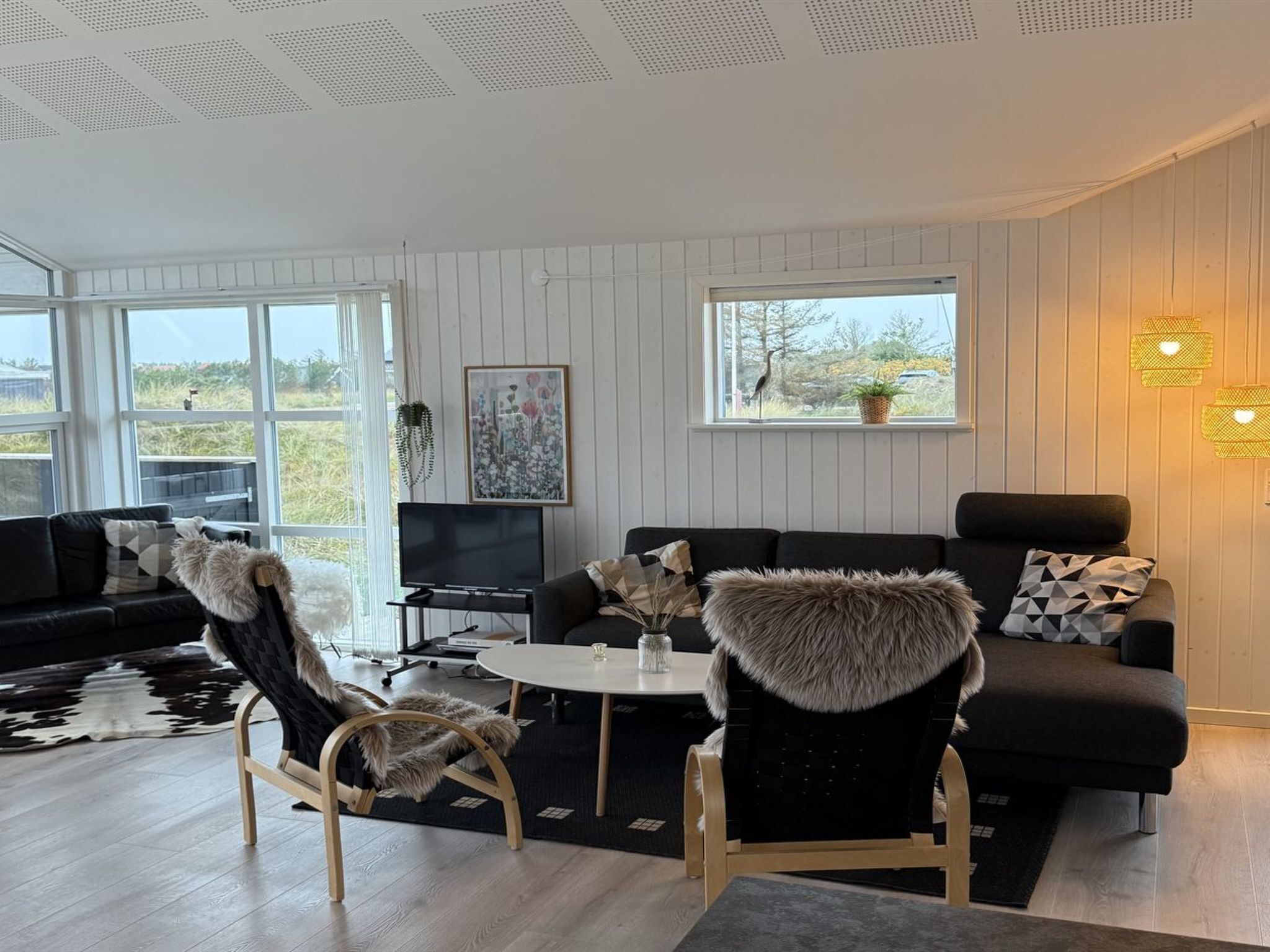 Ein modernes Wohnzimmer mit einer schwarzen Couch, zwei Sesseln, einem Couchtisch und einem Fernseher. Große Fenster bieten einen Blick ins Freie.