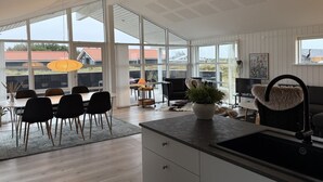 Et moderne køkken med en stor ø, et spiseområde med stole og bord, samt udsigt udendørs gennem store vinduer.