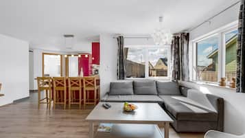 Un salon moderne avec un canapé d’angle, un coin repas avec une table et des chaises en bois, et une cuisine en arrière-plan.