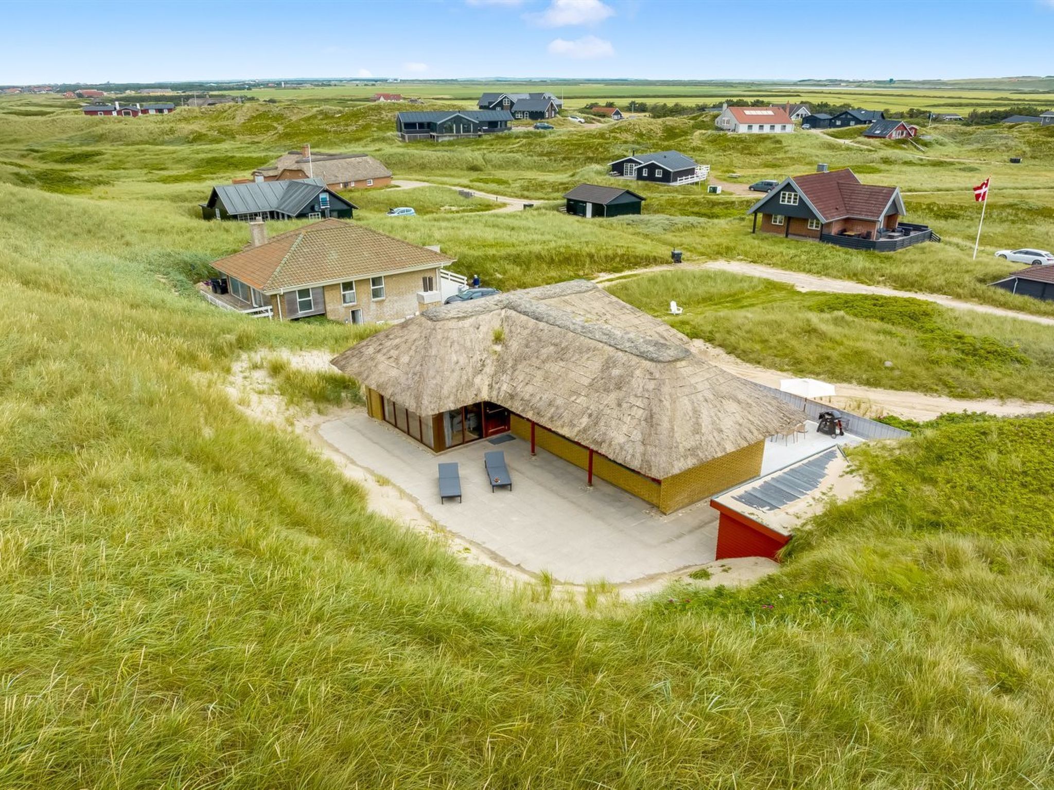 Een gebouw met rieten dak, een terras en lounge fauteuils, omgeven door grasvelden en andere huizen.