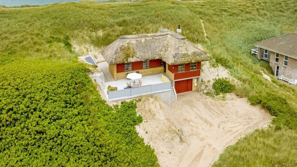 Une maison située sur la côte, avec un toit de chaume, une cour sablonneuse et une vue sur la mer et les éoliennes au loin.