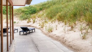 Une terrasse au bord de la plage, dotée d’un toit de chaume, de deux chaises longues et offrant une vue sur les dunes.