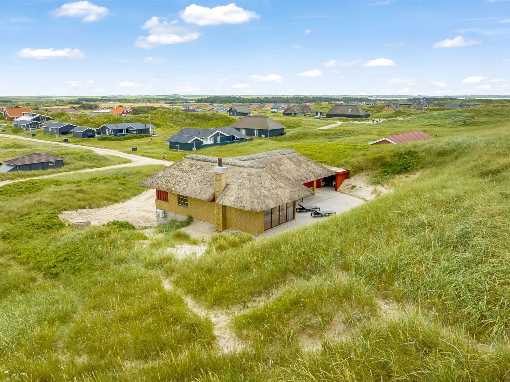 Ein Haus mit Strohdach, verbunden mit einem sandigen Pfad, umgeben von grasbewachsenen Dünen und weiteren ähnlichen Häusern im Hintergrund.