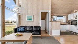 Ein modernes Interieur mit Holzbalkendecke, einem schwarzen Ledersofa, einem Holzesstisch und einer Küchenzeile mit weißen Schränken und Geräten.