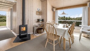 Ein modernes Holzinterieur mit einem zentralen Kamin, einem Essbereich mit Stühlen und einem Tisch sowie Blick durch große Fenster.