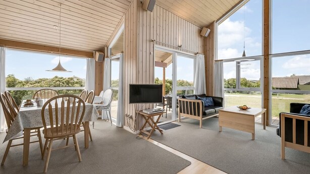 Ein geräumiger Raum mit Holzbalkendecke, großen Fenstern, einer Essecke mit Stühlen und Tisch, einem Sofa und einem Fernseher.