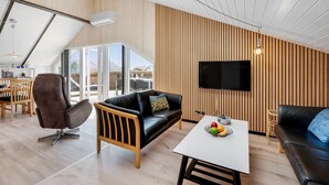 A modern living room with a wooden panel wall, a black leather sofa, a white coffee table with fruit, and a TV mounted on the wall. - "Arla" - 200m from the sea by Interhome (Harboøre)