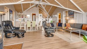 Een ruime woonkamer met hoge plafonds, een bank, twee leren fauteuils, een eettafel en een keuken op de achtergrond.