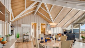 Een ruime loft met een hoge plafond, zichtbare houten balken, een eethoek met tafel en stoelen, en uitzicht op het omliggende landschap.