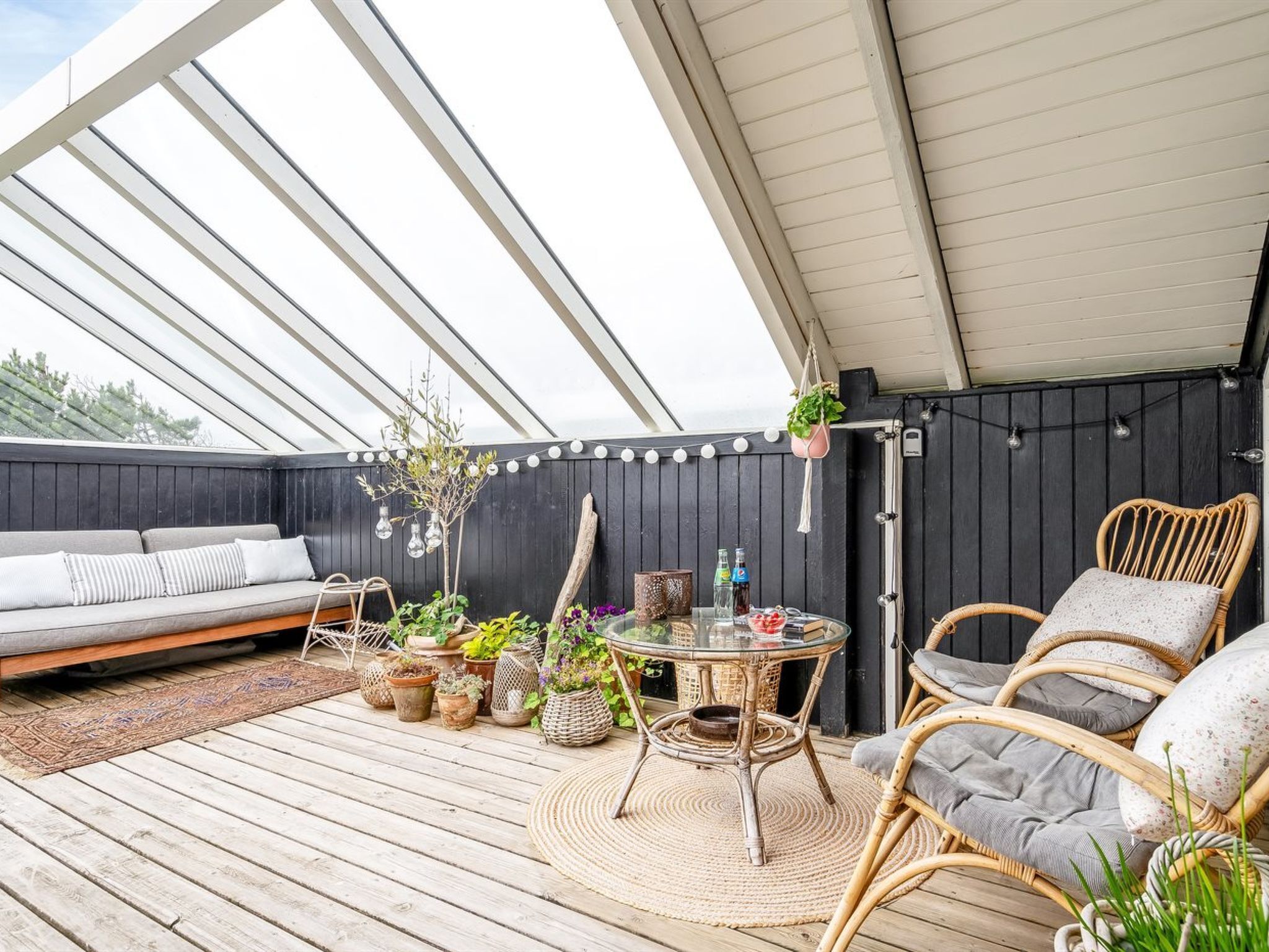 An outdoor patio with a wooden deck, a round table, wicker chairs, a sofa, and various potted plants.