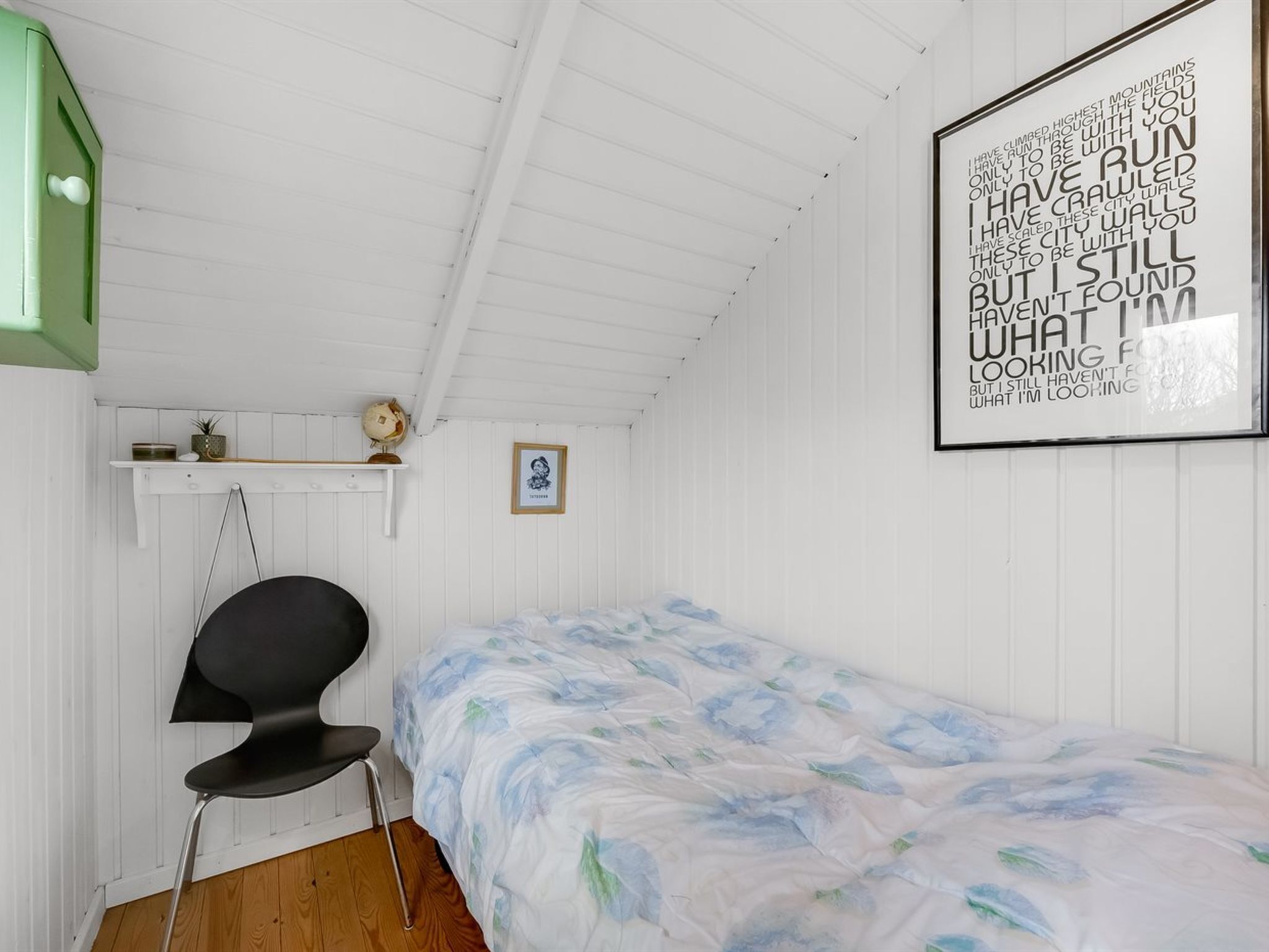 A small, single-bed room with a white ceiling and walls, a black chair, a bed with a floral bedspread, a shelf with decorative items, and a motivational quote on the wall.