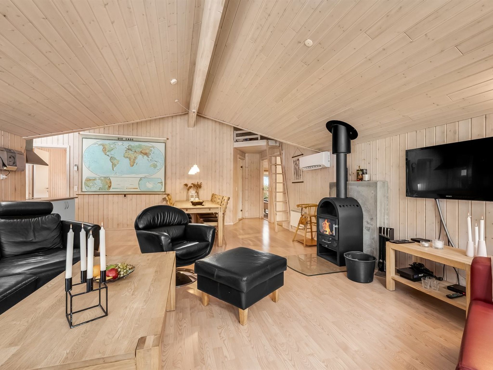 Een gezellige woonkamer met een houten plafond, een zwarte leren bank, een houten eettafel, een zwart leren poef, een zwart houtkacheltje, een televisie en een kaart aan de muur.