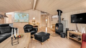 Una sala de estar acogedora con techo de madera, un sofá de cuero negro, una mesa de comedor de madera, un reposapiés de cuero negro, un estufa de leña negra, un televisor y un mapa en la pared.