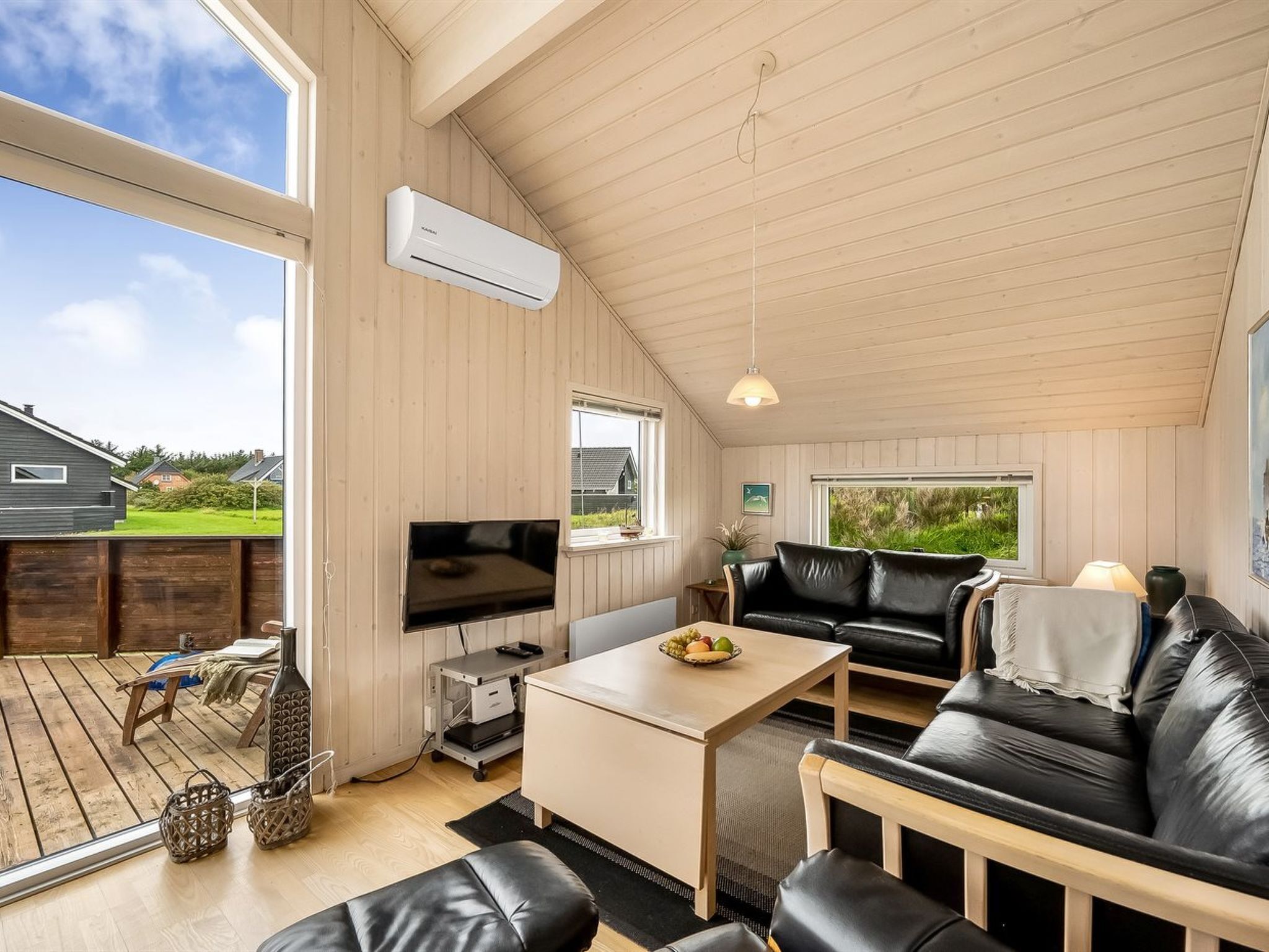 Un vaste espace de vie doté d’une grande fenêtre, d’un téléviseur à écran plat, d’un canapé en cuir, d’une table basse et d’une terrasse en bois visible à travers la vitre.