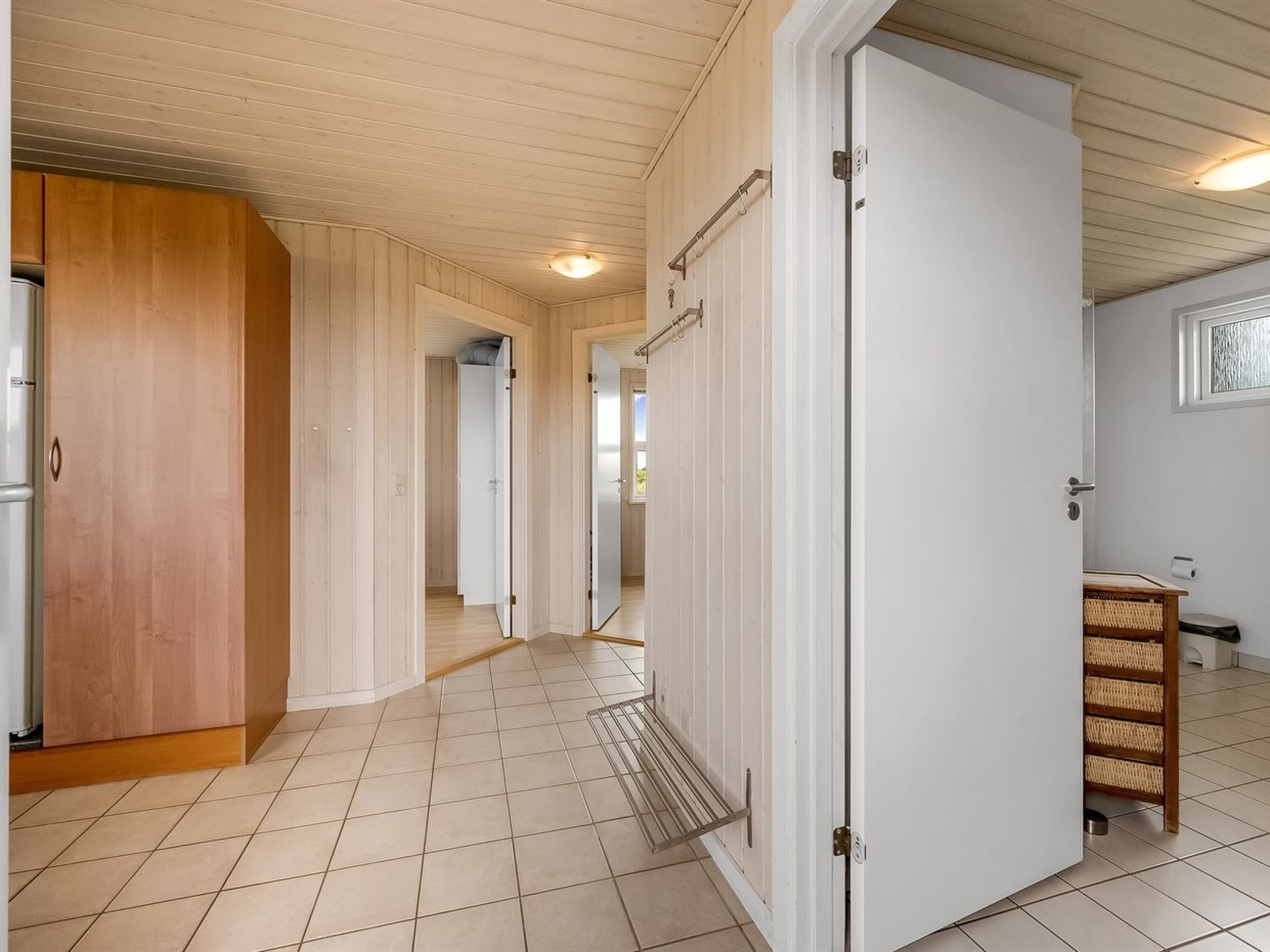 Een gang met houten wanden en een betegelde vloer, die leidt naar een kamer met een witte deur en een klein plankje met manden.
