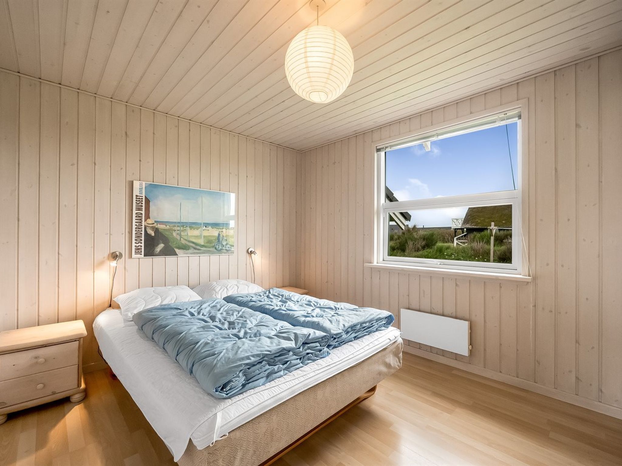 A bedroom with a bed, a nightstand, a wall-mounted lamp, a window with a view, and a poster on the wall.