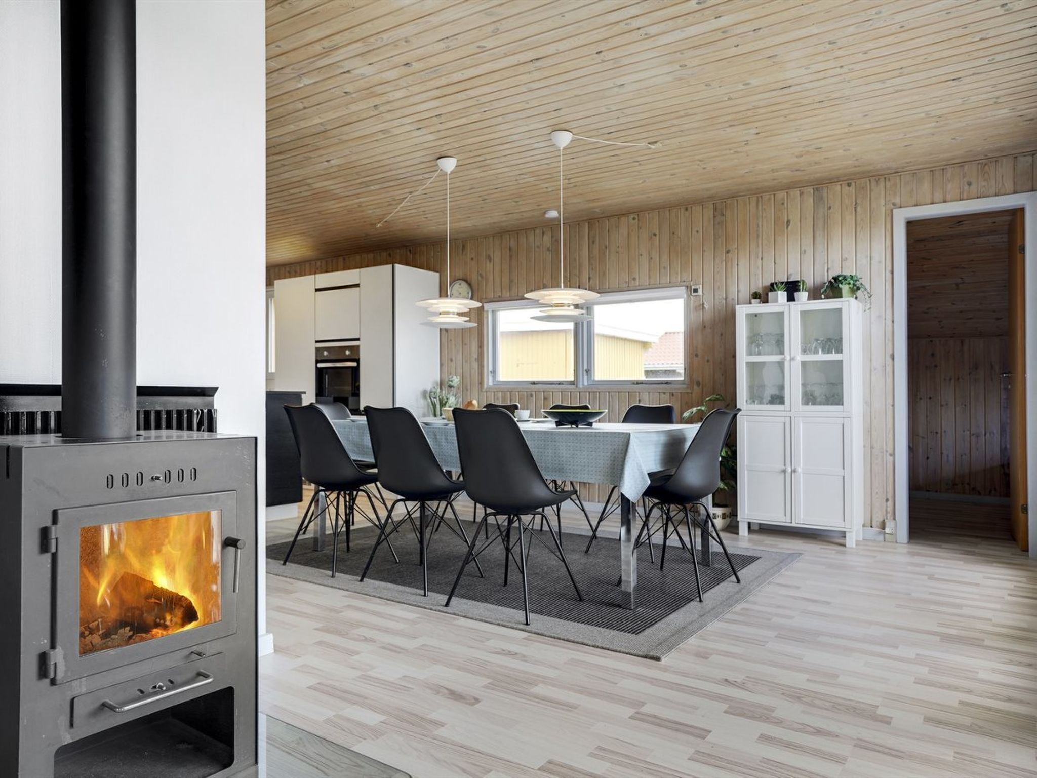 A modern kitchen with a wood-burning stove, dining area with chairs, and a view of the outside through large windows.