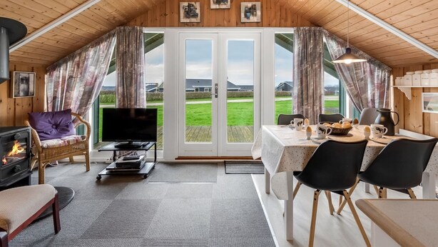 Een gezellig interieur met een houten plafond, een open haard, een televisie, een eethoek en grote ramen met uitzicht op de omgeving.
