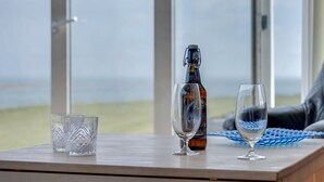 Et bord med en flaske og to glas, en blå vævet dug og udsigt til stranden gennem store vinduer.