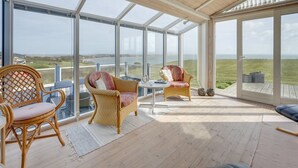 Et rummeligt solrum med store vinduer, der giver udsigt over havet og et græsslået landskab.
