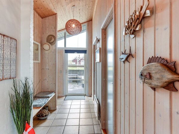 A wooden hallway with a tiled floor, a hanging light fixture, a door leading outside, and wall decorations including a fish and wooden tools.