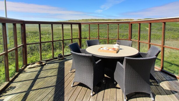 A wooden deck with a round table and wicker chairs, overlooking a grassy landscape.