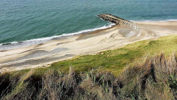 Paisagem costeira com uma praia arenosa, um molhe rochoso e dunas gramadas.