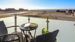 Varanda com cadeiras de vime e uma mesa pequena, com vista para uma praia com casas e uma estrada.