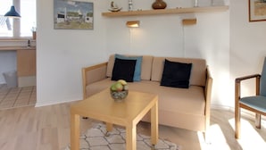 Uma sala de estar com um sofá bege, uma mesa de centro de madeira e um tapete pequeno. Na parte de trás, há uma cozinha com uma janela e um lustre.