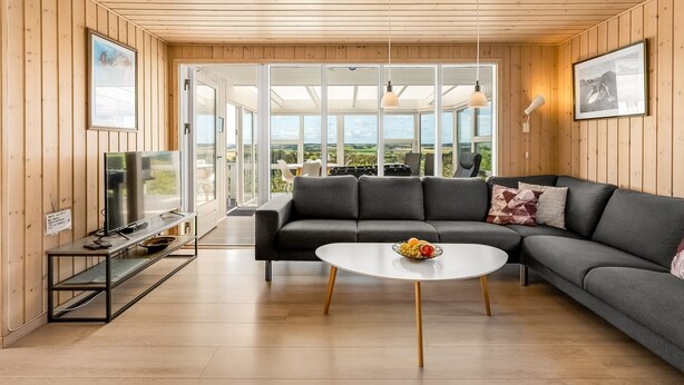 Ein modernes Wohnzimmer mit einer Eckcouch, einem runden Couchtisch und einem Fernseher. Große Fenster bieten einen Ausblick ins Freie.