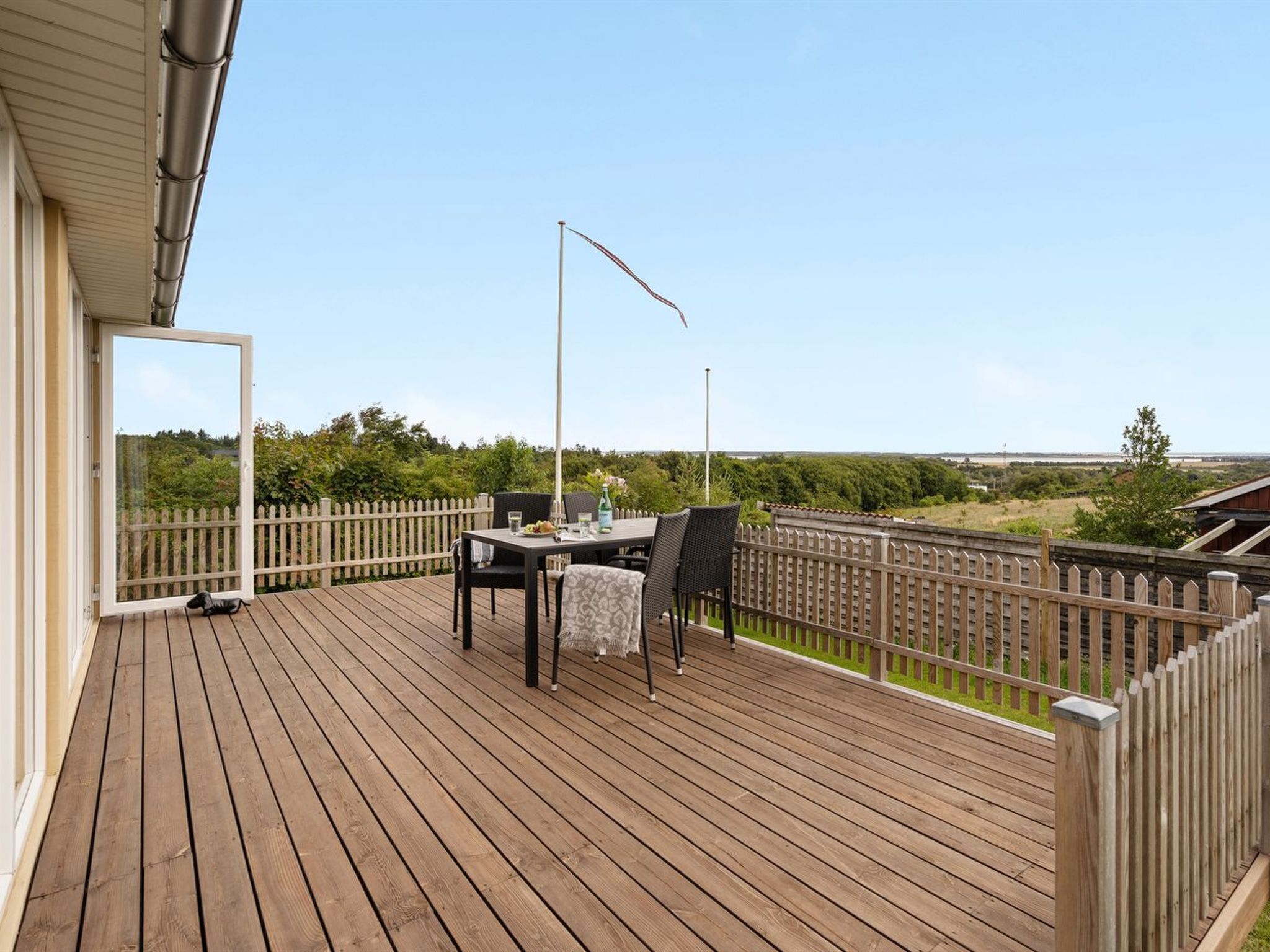 Een houten terras met een tafel en stoelen, een metalen hekwerk en uitzicht op het platteland.