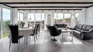 A spacious, modern living area with a dining table, chairs, a sofa, and a large window offering a view of the outdoors.
