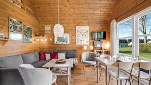 Een gezellig cabinteinterieur met een houten plafond, een grijze bank, een eettafel en een aan de muur bevestigde televisie.